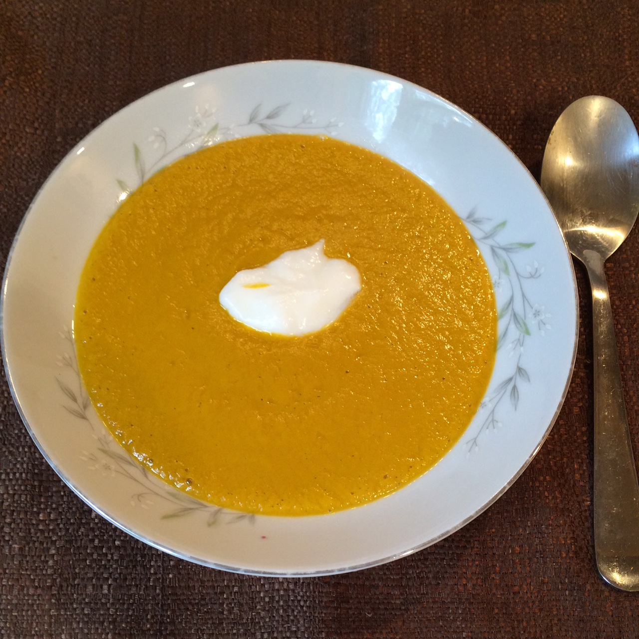 Low profile white china bowl with a deep orange soup and small dollop of white goat yoghurt in the center, silver spoon on the side, centered on a brown fabric background.