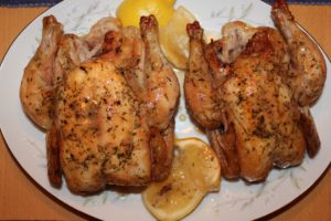 Two Cornish game hens, side by side, one a white china plate, with herbs on top and two lemon halves.