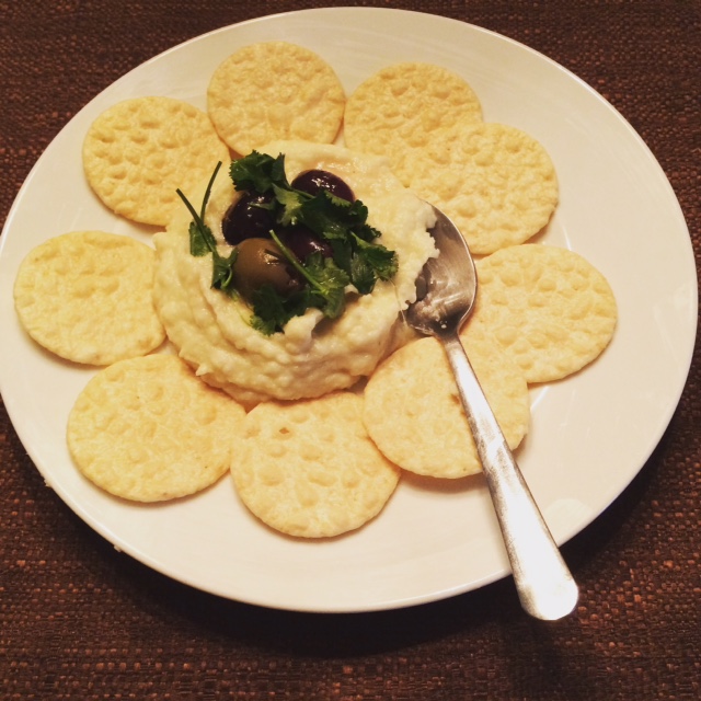White plate on brown background, round rice crackers surrounding full cup scoop of cauliflower hummus, topped with parsley and four Greek olives, silver spoon on side of hummus. 