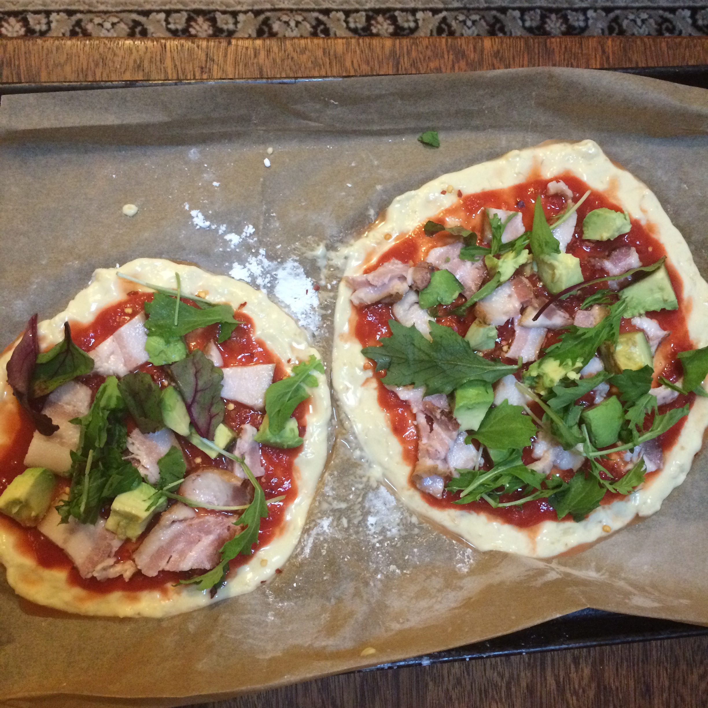 Two small pizza crusts with bacon pieces, leafy vegetables, avocado cubes, and diced tomatoes in sauce, laying on parchment paper before baking.