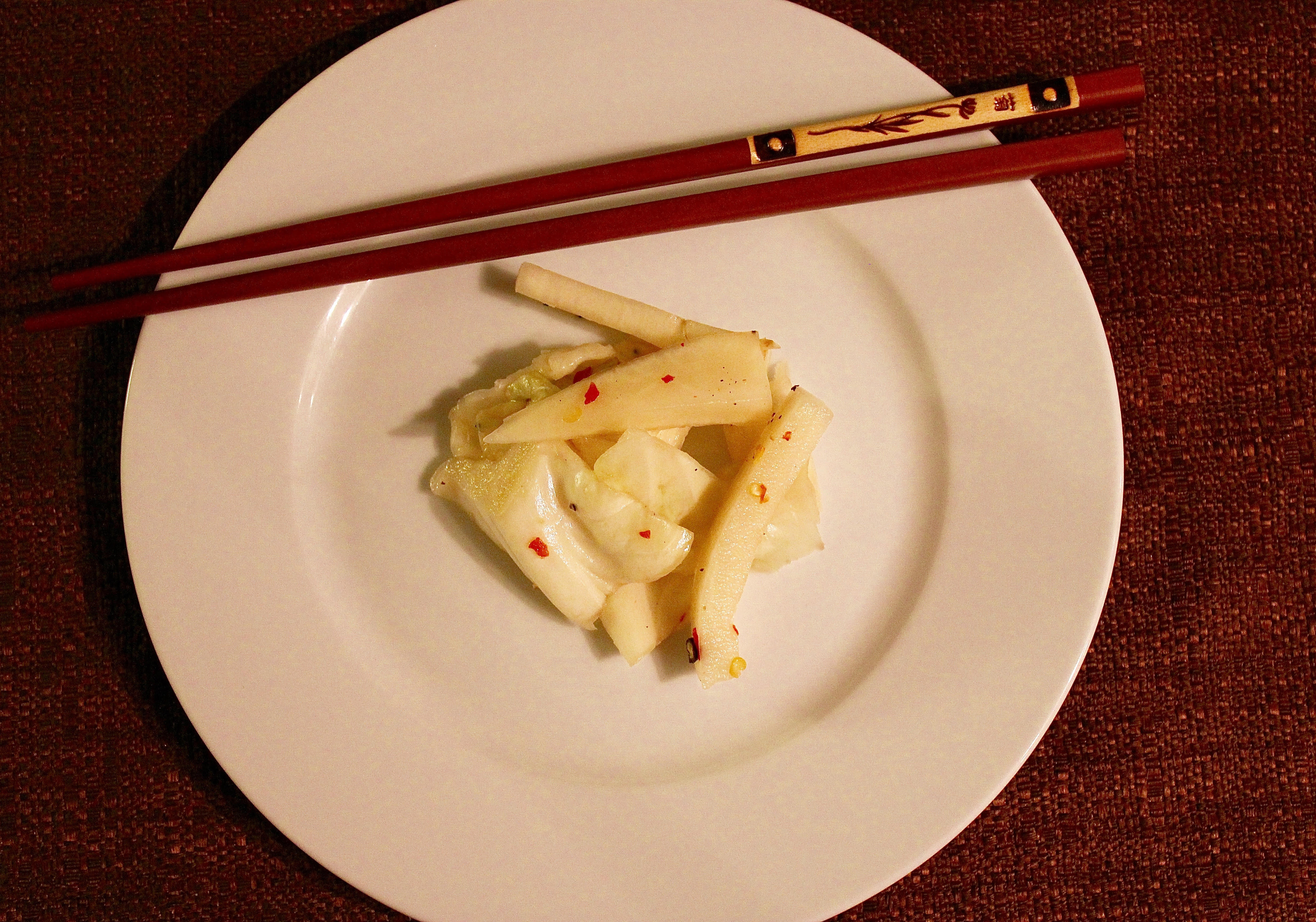Dikon vegetable and cabbage, after pickling, in the center of a white plate on a brown background, with a pair of deep red chopsticks across the top of the plate.