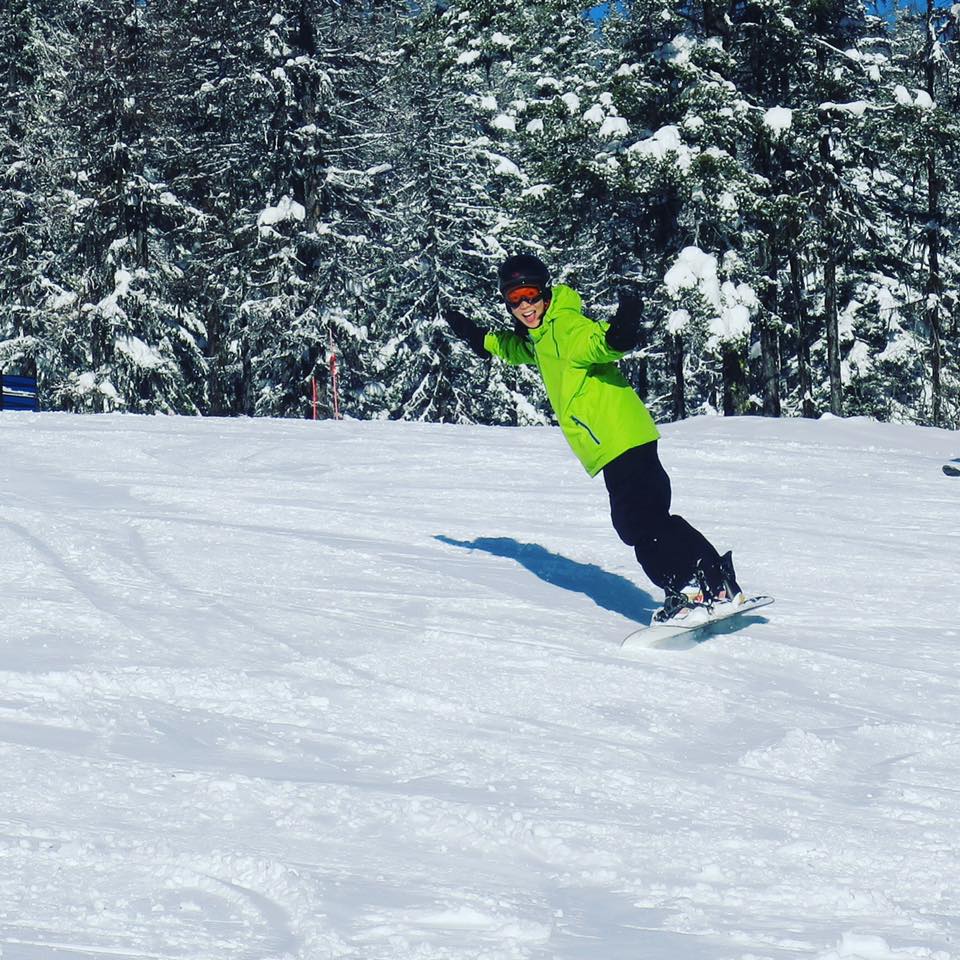 Author, wearing a bright green snowboard jacket and black pants, helmet and goggles, boarding down a snow-covered hill. 