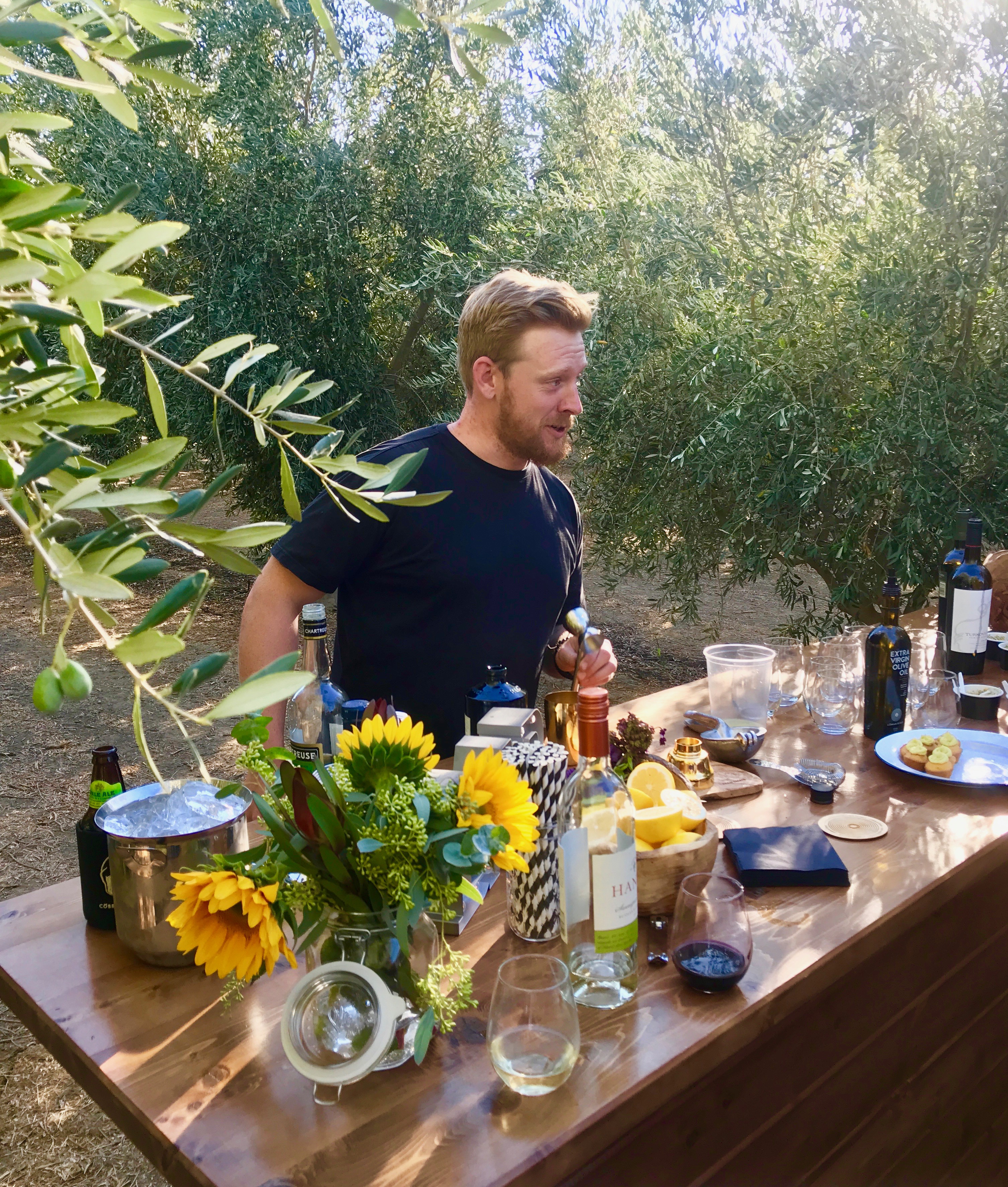 Chef with beard stands behind counter in the middle of an olive grove. Table has wine, steel drink container, flowers, and small plates of food.
