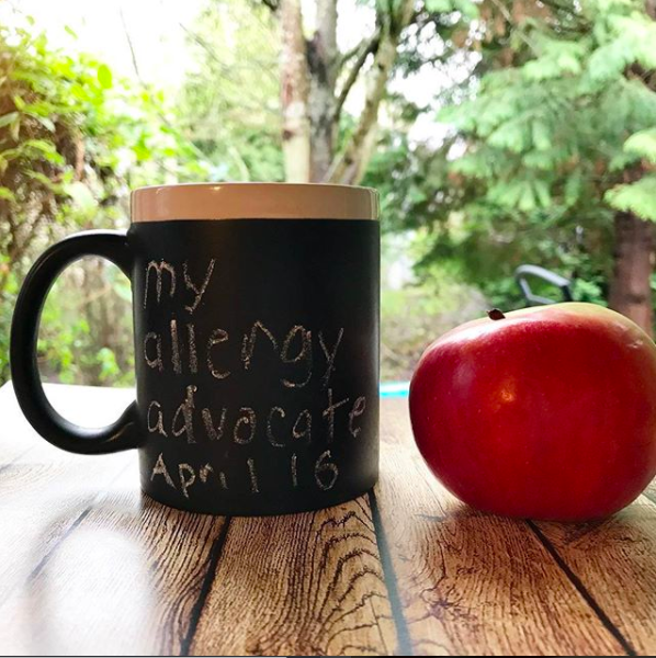 Black mug with words, "My Allergy Advocate April 16" in chalk, apple next to cup, resting on a wood board, trees in the background of morning light